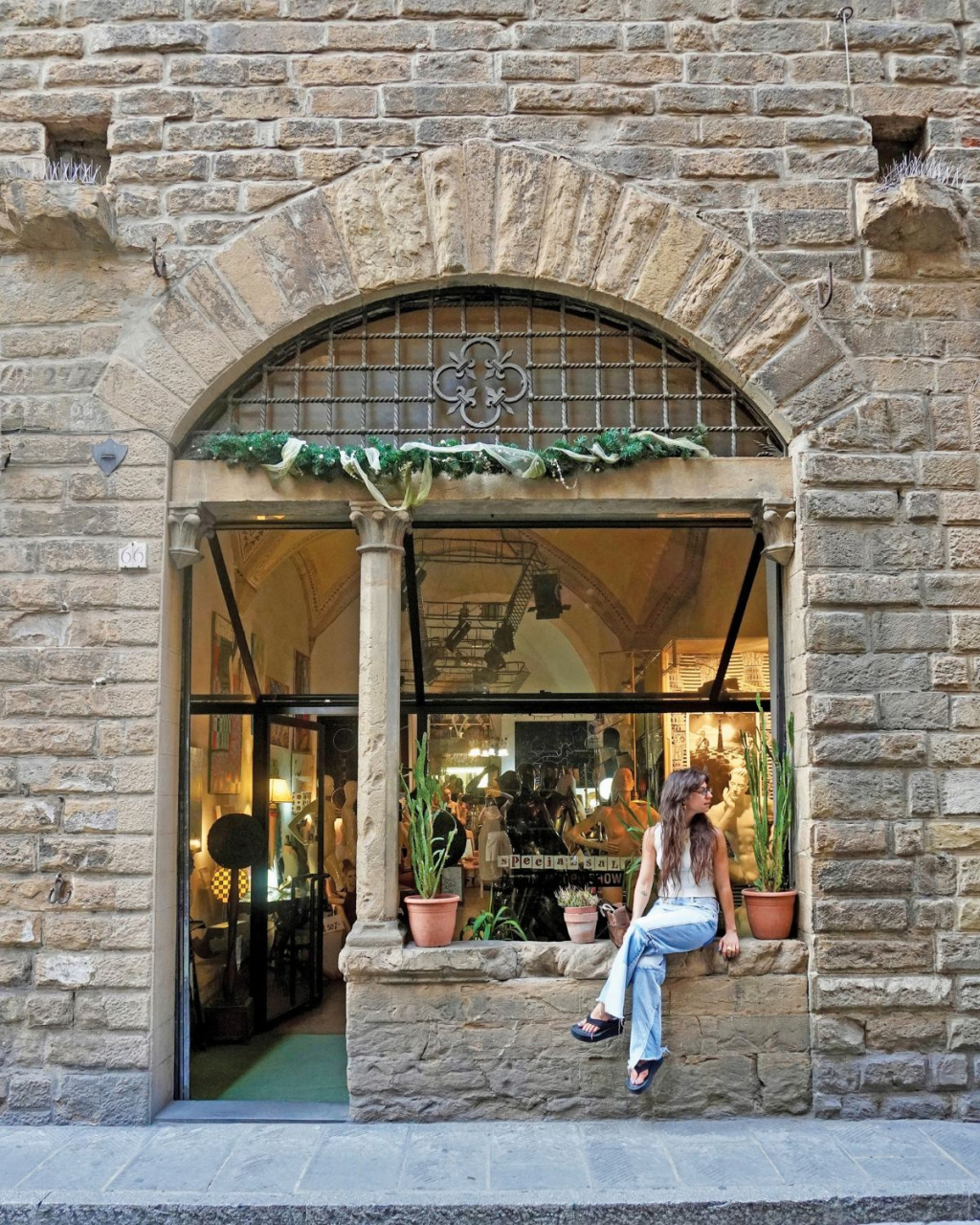 A woman in jeans sits on a stone window ledge outside a warmly lit shop, framed by an arched brick façade, potted plants, and greenery.