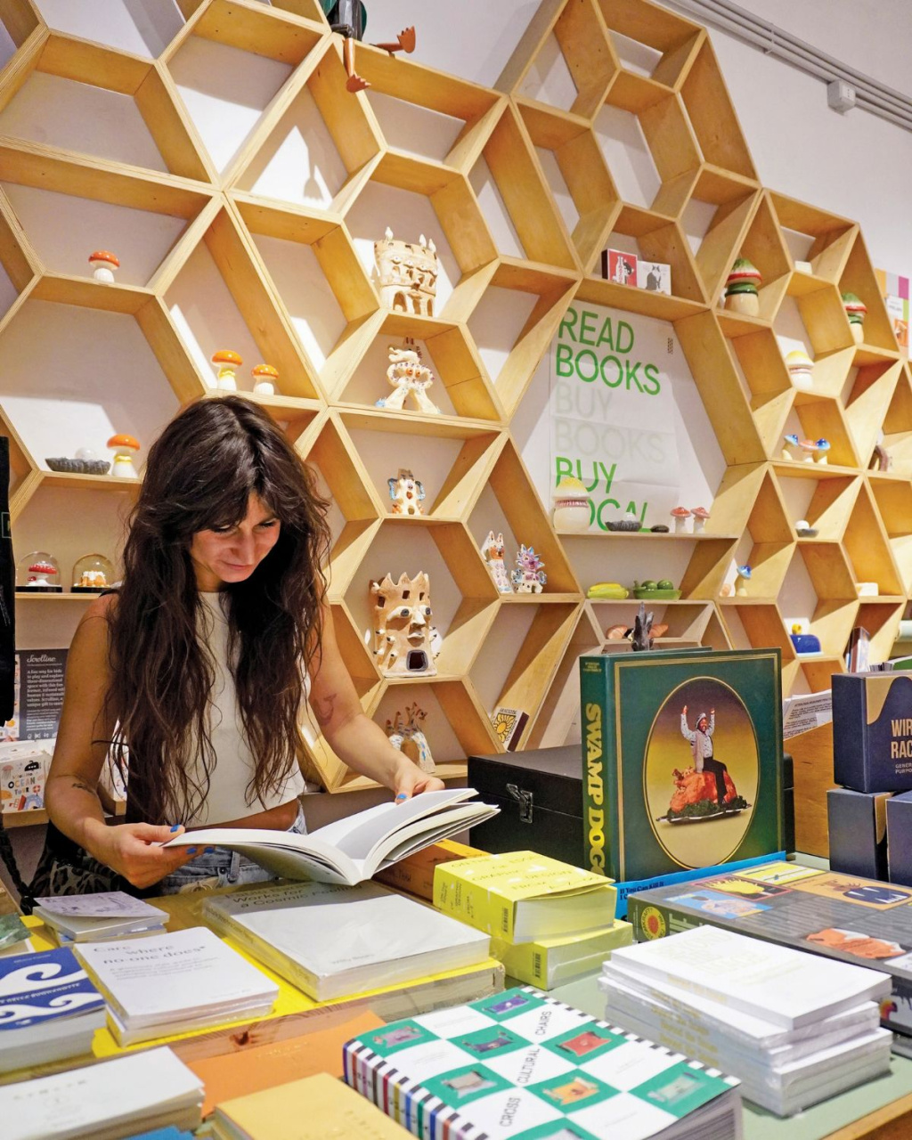 A woman browses an open book at a colorful bookstore counter, surrounded by stacked books, small ornaments, and honeycomb shelves.