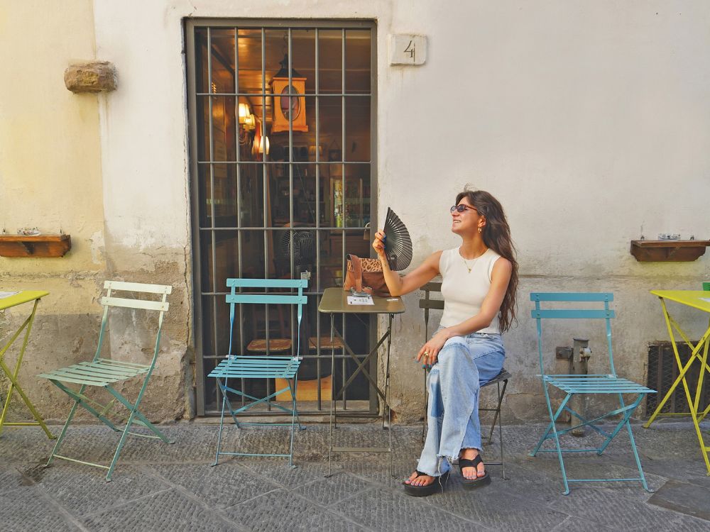 A woman in sunglasses sits outside a café, fanning herself beside colorful metal bistro chairs; warm light gives the quiet street a relaxed mood.