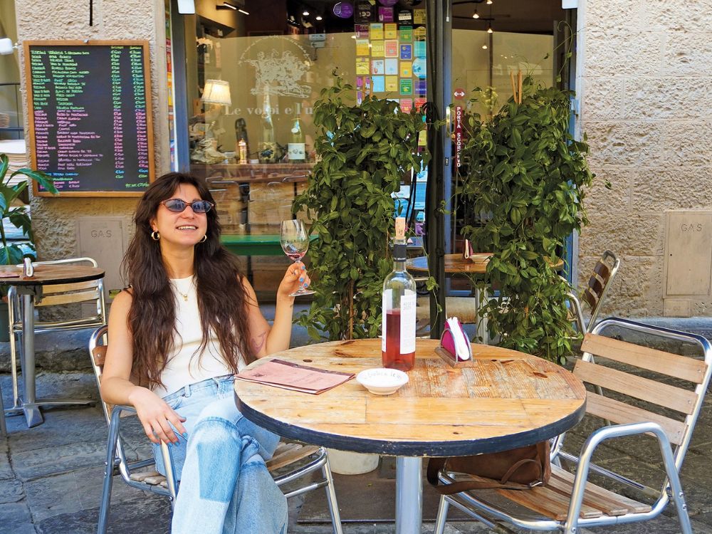 A smiling woman in sunglasses relaxes at a sidewalk café, holding a wine glass beside a bottle on a wooden table amid leafy plants.