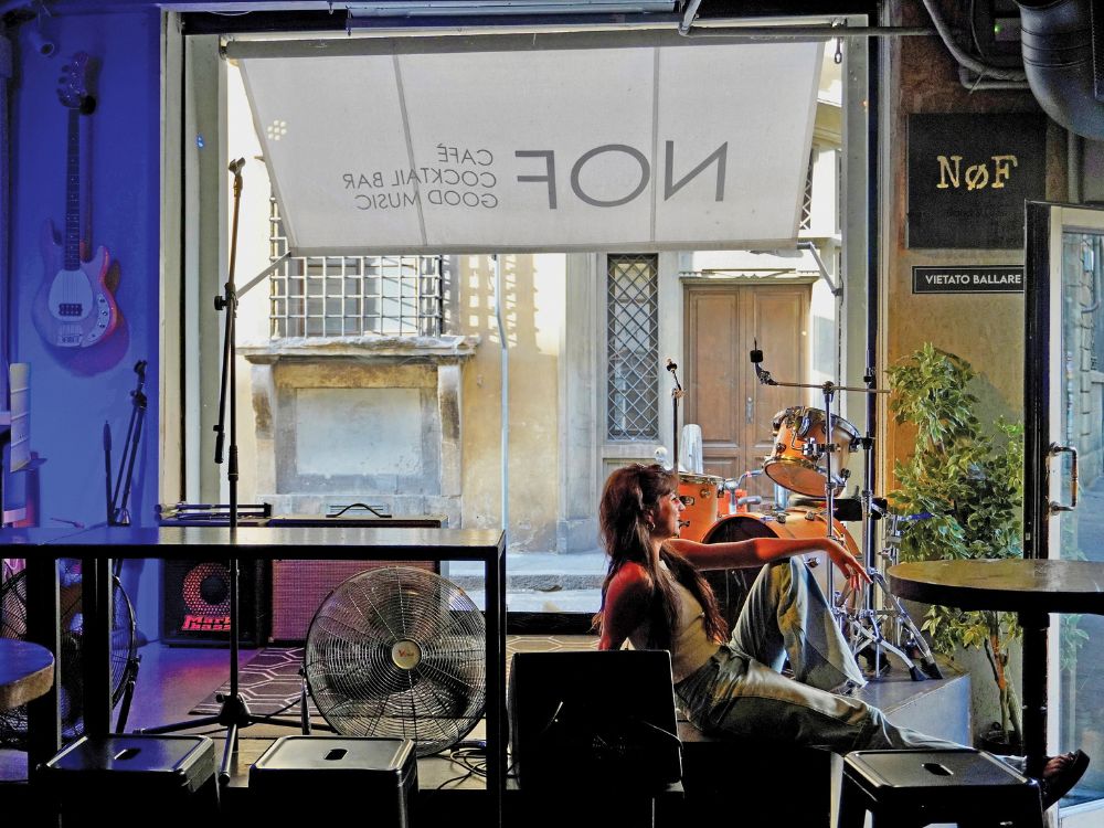 A relaxed musician sits with a guitar by a café window, surrounded by drums and a floor fan; colorful lighting gives the room an intimate, creative mood.