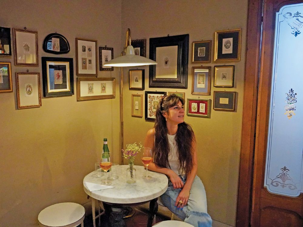 A woman sits in a cozy vintage café, surrounded by framed art and a marble table with drinks and flowers, creating a warm, nostalgic mood.