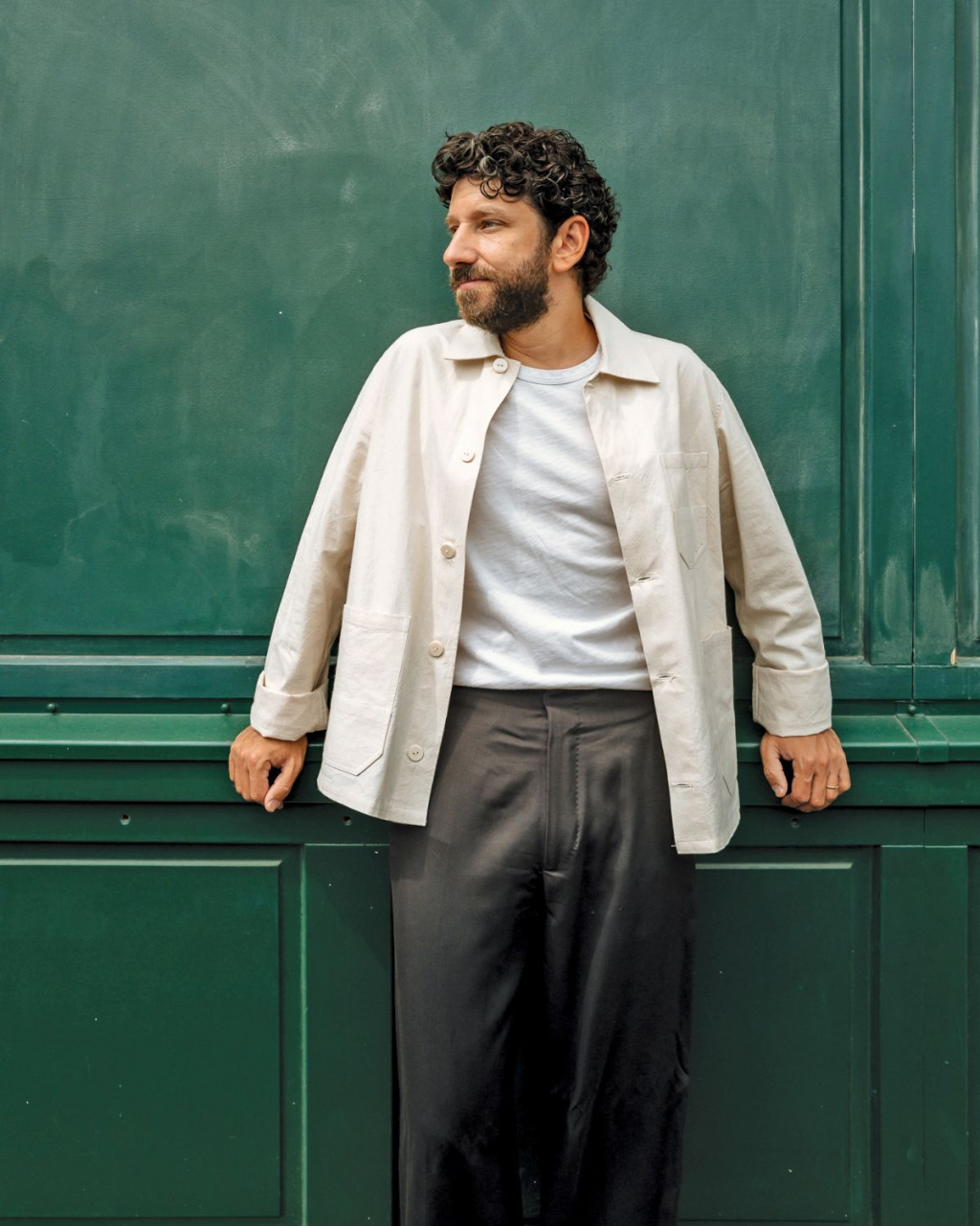 A curly-haired, bearded man (Ilan Molcho) in a cream jacket, white shirt, and dark trousers leans against a green paneled wall.