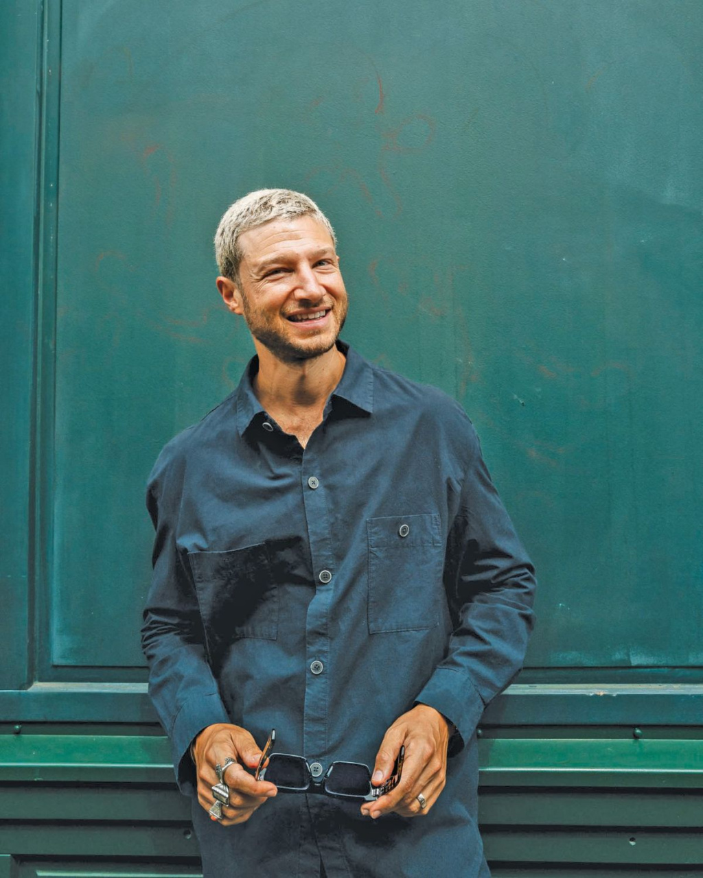 A smiling man with short silver hair (Nuriel Molcho) stands before a teal-green wall, holding sunglasses in both hands.