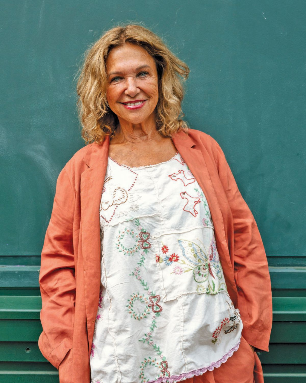 Smiling woman with wavy blonde hair (Haya Molcho) poses before a green wall, wearing a coral jacket and floral embroidered top.