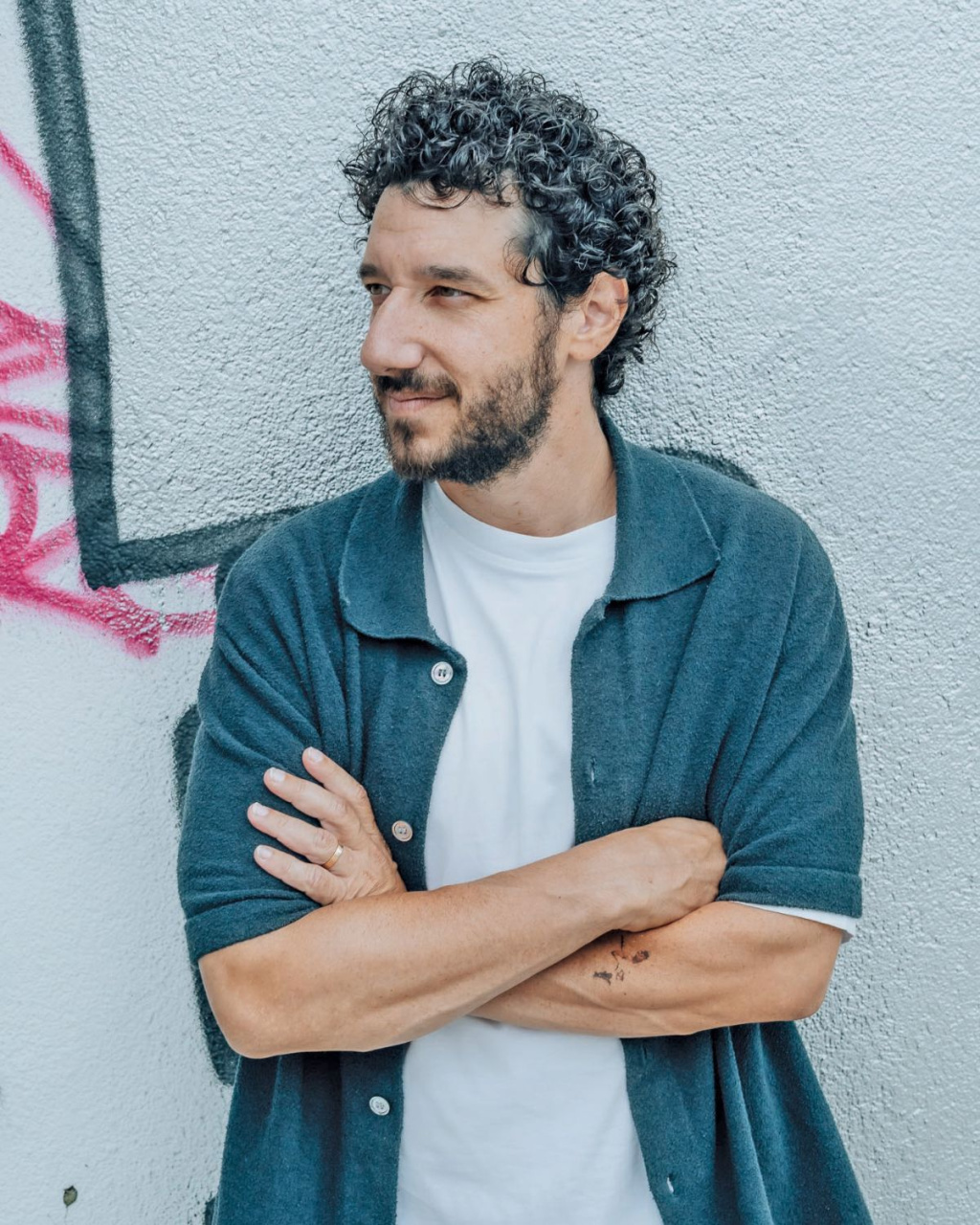 A curly-haired, bearded man (Elior Molcho) in a teal cardigan stands with arms crossed against a graffiti-marked concrete wall.